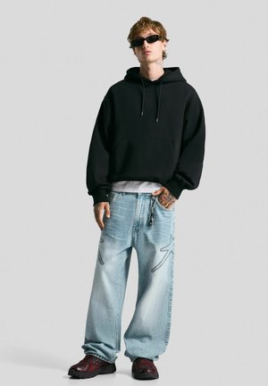 Black hooded sweatshirt, light blue wide-leg jeans with stitching accents, and burgundy sneakers. Accessories include sunglasses and keychain.
