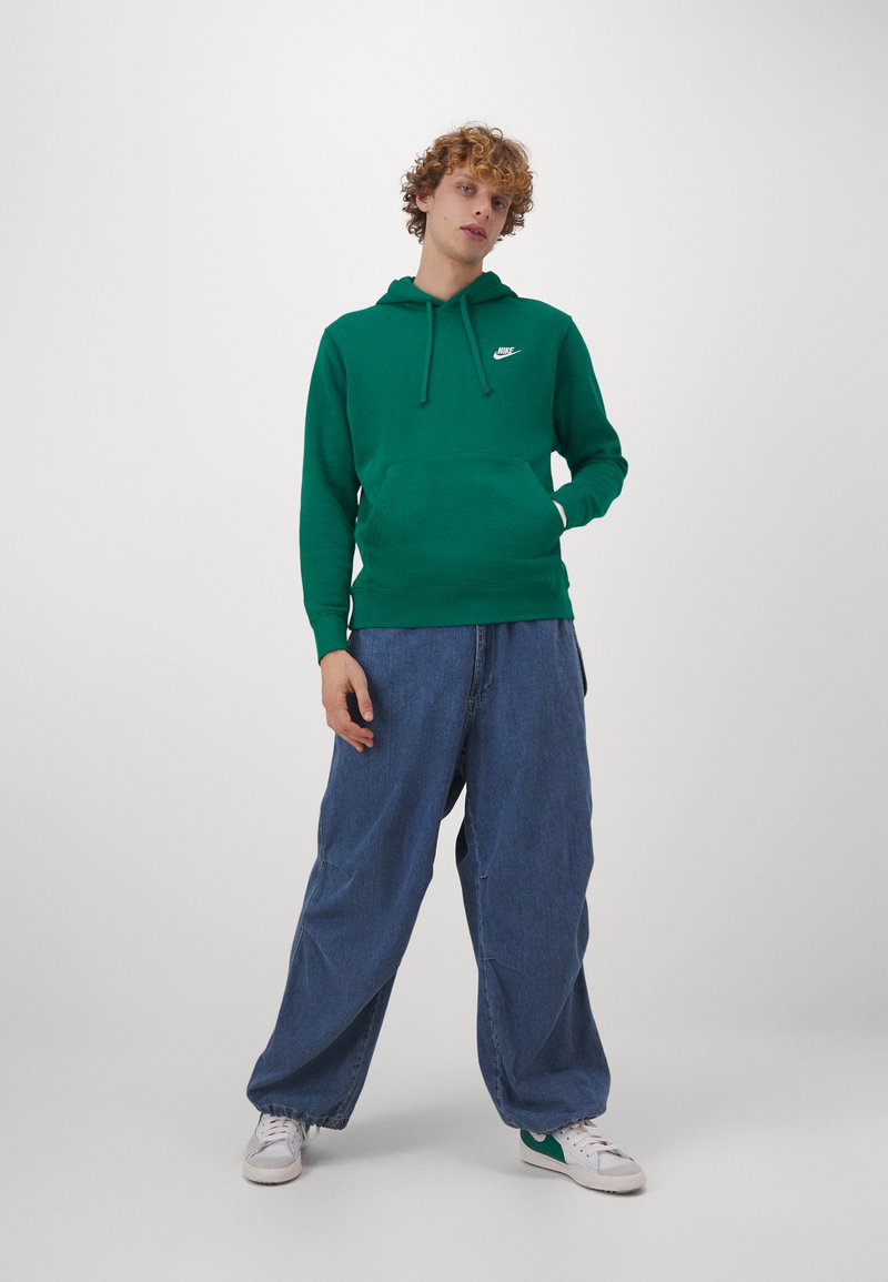 Sudadera de algodón verde con bolsillo frontal y capucha ajustable con cordón, combinada con unos vaqueros de mezclilla azul de corte holgado y zapatillas blancas.