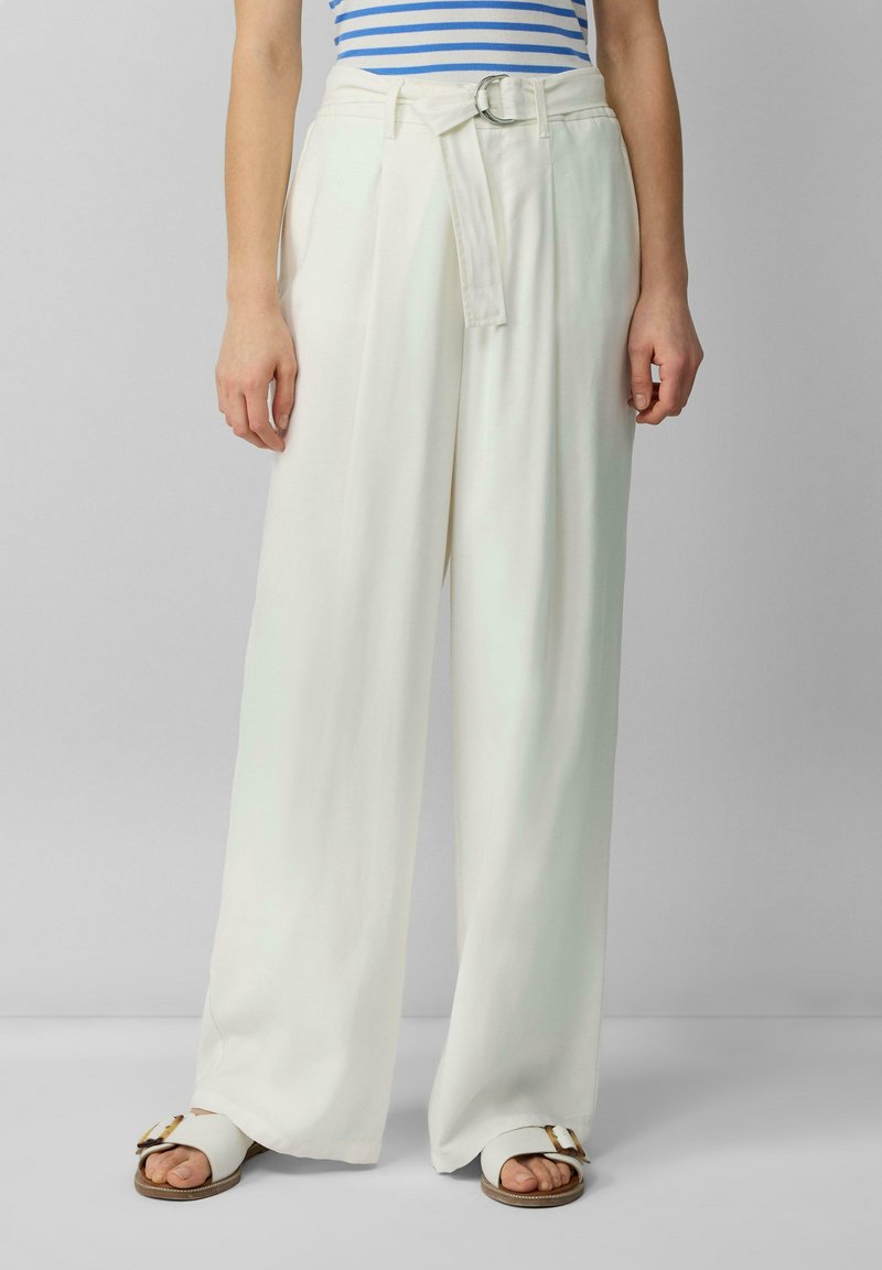 Witte wijde broek met een stoffen riem, voorzien van een cirkelvormige gesp. Glad textuur met een gepaste pasvorm, gecombineerd met platte sandalen.