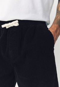 Zwarte corduroybroek met geribbelde textuur, voorzien van een elastische tailleband en een wit trekkoord. Zakjes zijn zichtbaar aan weerszijden.