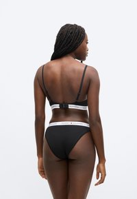 Černá podprsenka a kalhotky v sadě s tenkými ramínky. Podprsenka má bílý proužek s logem značky. Materiál je hladký; design je minimalistický.