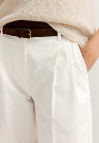 White pleated trousers with side pockets, paired with a beige knit top. A brown suede belt with a gold buckle accents the outfit.
