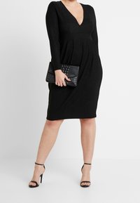 Czarna, długa sukienka z długim rękawem i dekoltem w serek, z dopasowanym gorsetem i rozkloszowaną spódnicą. Połączona z teksturowaną czarną kopertówką i sandałami na kostkę.