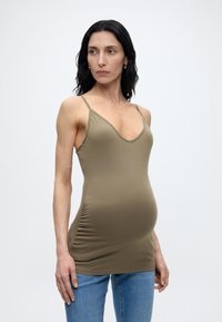 Έγκυος γυναίκα με μαύρα μαλλιά φοράει αμάνικο τοπ ελιάς και μπλε τζιν, στέκεται μπροστά σε απλό λευκό φόντο.