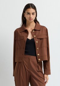 Kvinna med långt mörkt hår som bär guldörhängen, brun jacka med knappar och stora fickor, samt matchande bruna byxor över en svart topp.