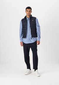 Marine gewatteerde vest over een lichtblauw en wit gestreept langarmshirt, gecombineerd met marineblauwe broek en witte sneakers. Gladde stof, gestikt ontwerp.