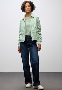 Giacca in corduroy verde menta con due tasche frontali, abbinata a un top verde chiaro e jeans a gamba larga blu scuro. Sneakers bianche completano l'outfit.