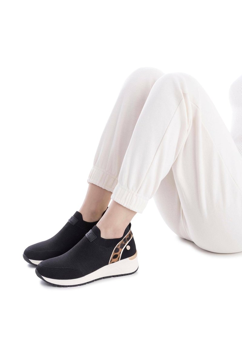 Zapatillas negras sin cordones con un upper texturizado, acento de estampado de leopardo en el talón y una suela blanca ligera, combinadas con joggers blancos de cintura elástica.