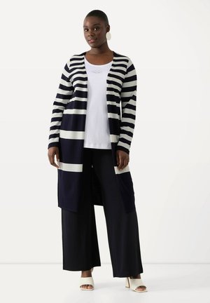 Gestreepte lange cardigan in marineblauw en wit, gecombineerd met een witte top en brede zwarte broek. Slanker breisel, open voorkant, zijzakken.