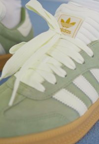 Zaļas zamšādas kedas ar baltu gumijas zoli, trim baltām svītrām un dzelteniem šņorēm. Adidas logo uz brūnas etiķetes netālu no valkāšanas daļas.