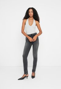 Sivi halter bodi u kombinaciji s tamnosivim skinny trapericama i crnim cipelama na visoku petu s oštrim vrhom. Glatke teksture i oblikovanje koje se prilagođava tijelu.
