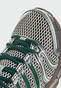 Athletic shoe featuring a mesh upper in silver and green, synthetic accents, and a textured brown rubber sole. Notable lacing system design.