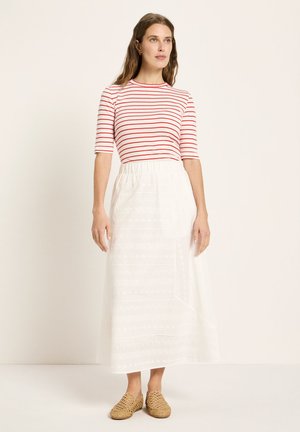 Frau steht vor einem einfarbigen Hintergrund, trägt ein rot-weiß gestreiftes Oberteil, einen weißen knöchellangen Rock und geflochtene beige Slipper.