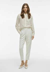 Blusa larga de manga larga blanca y texturizada con botones al frente, combinada con pantalones crema ajustados y tacones blancos de punta.