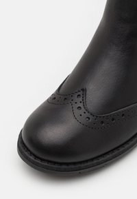 Bota de cuero negro con punta redondeada, que presenta un detallado de adorno tipo brogue y una suela cosida. Textura suave y acentos de diseño mínimos.