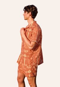 Camicia a maniche corte e pantaloni coordinati in corallo con motivi botanici crema. Tessuto leggero, vestibilità rilassata e colletto con bottoni.
