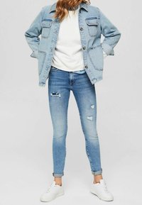 Chaqueta de denim azul claro con dos bolsillos en el pecho, llevada sobre un suéter blanco. Jeans skinny azules desgastados y rasgados con puños doblados y zapatillas blancas.