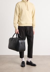 Bolsa de deporte de cuero negro con asas dobles, un zipper plateado y forma redondeada. Llevada con una sudadera amarilla clara y pantalones negros.
