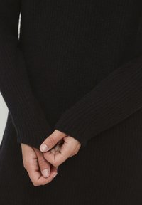 Pullover nero in maglia con texture a coste, con maniche lunghe. Mani unite, che mettono in evidenza il tessuto morbido e i polsini.