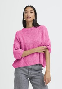Femme portant un pull en tricot surdimensionné rose vif et un jean gris, ajustant une manche, avec de longs cheveux foncés et des boucles d'oreilles en argent.