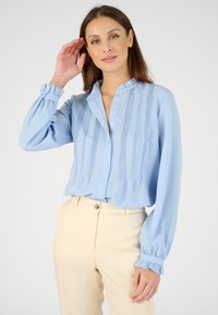 Blusa de botón azul claro con pliegues verticales, cuello con volantes y puños elásticos, combinada con pantalones beige. Textura de tela suave.