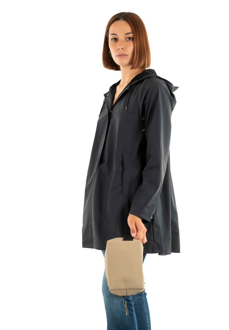 Mørk marineblå regnjakke med hette, med glidelåslommer og glatt tekstur. Beige kompakt veske holdt i venstre hånd.