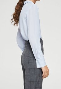 Persona con una blusa a maniche lunghe azzurro chiaro infilata in pantaloni grigi a quadri a vita alta, mostrata di lato con lunghi capelli ricci.
