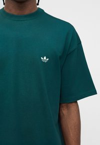 Chemise bleu sarcelle foncé à manches courtes avec petit logo blanc Adidas trefoil sur le côté gauche de la poitrine, portée par une personne à la peau foncée et aux cheveux tressés courts.