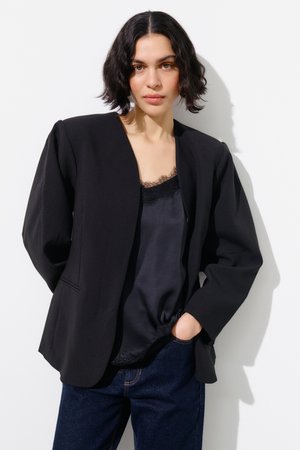 Kvinde med kort bølget mørkt hår iført en sort blazer, sort camisole med blonder og mørkeblå jeans, stående mod en hvid baggrund.