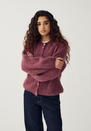 Kobieta o długich, kręconych włosach, ubrana w miękki, fioletowy kardigan na guziki oraz ciemne dżinsy, stojąca z założonymi ramionami na jednolitym tle.