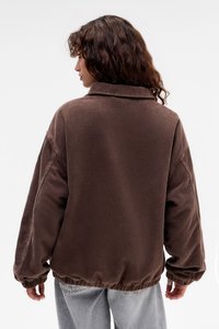 Mujer con cabello ondulado que lleva una chaqueta de pana marrón oversized y jeans de color azul claro, de espaldas frente a un fondo blanco liso.