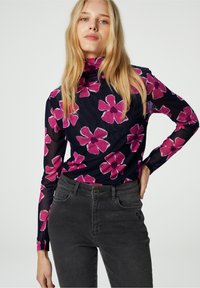 Langmouwige, doorzichtige top met een col, marineblauwe basis en een groot roze bloemenpatroon. Gepaard met donkergrijze jeans met een knoopsluiting.
