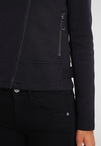 Černá, slim fit bunda s kapsou na zip a texturovanou látkou. Má vysoký límec a štíhlý design, doplněnou černými džíny.