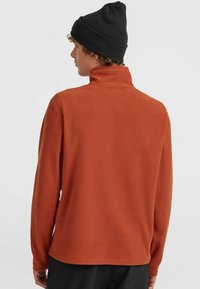 Fleecepullover i bränd orange, med hög krage och långa ärmar. Materialet har en mjuk textur, med en rak fåll längst ner.