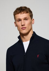 Marinblå zip-up-tröja med hög krage, gjord av ett stickat material, med en liten röd logotyp på vänster bröst.