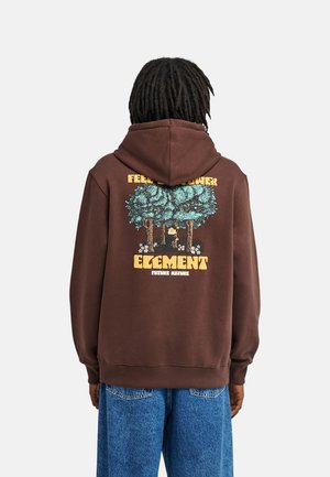 Persona con dreadlocks scuri indossa una felpa marrone con grafica forestale e la scritta ""Feel The Power Element Future Nature"" sulla schiena, e jeans blu.