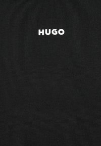 Țesătură tricotată neagră cu numele brandului "HUGO" scris cu litere albe îndrăznețe, poziționat proeminent. Textură moale, fără alte modele.