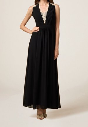 Czarna sukienka maxi bez rękawów z koronkowym dekoltem w szpic i plisowanym górnym dołem, noszona z beżowymi sandałami na otwartym palcu, modelka z dłonią na biodrze.