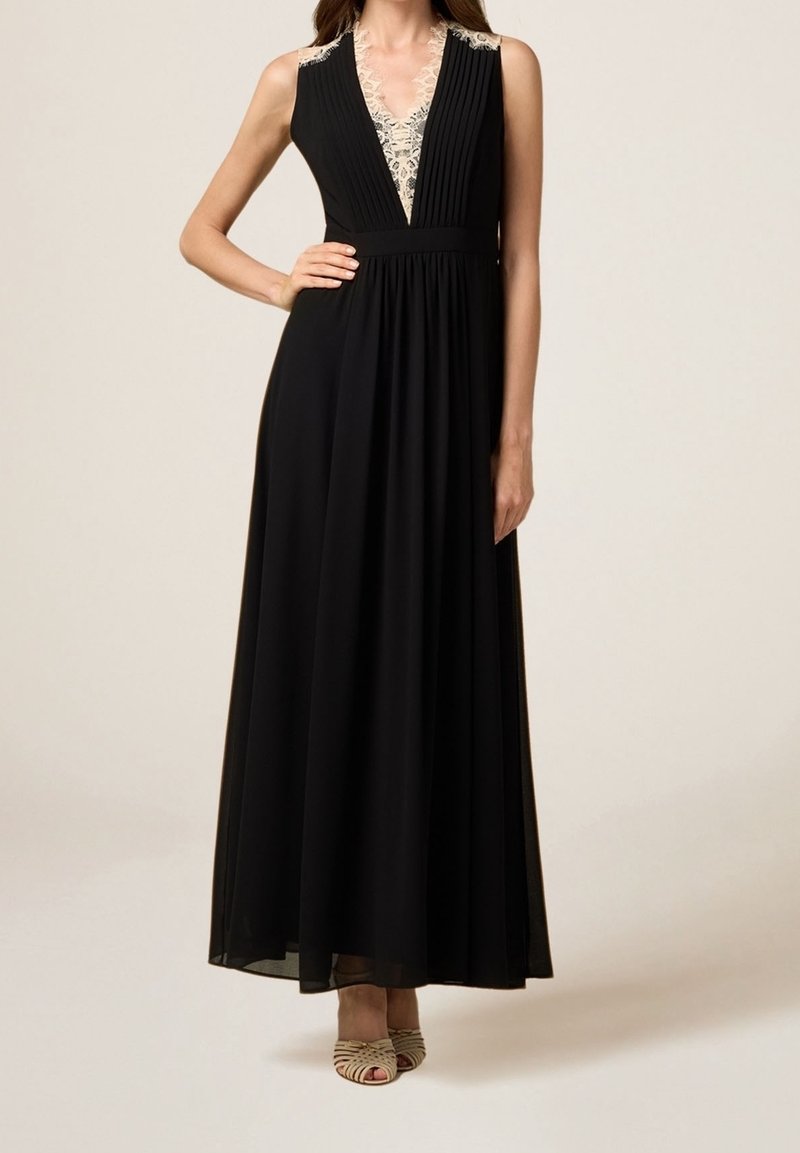 Robe maxi noire sans manches avec détail en dentelle sur le col en V et corsage plissé, portée avec des talons ouverts beige, mannequin main sur la taille.