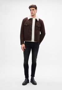 Hnedá semišová bunda s bielym kožušinovým golierom a dvoma náprsnými vreckami, nosená cez krémový sveter a čierne slim-fit džínsy. Čierne členkové čižmy.