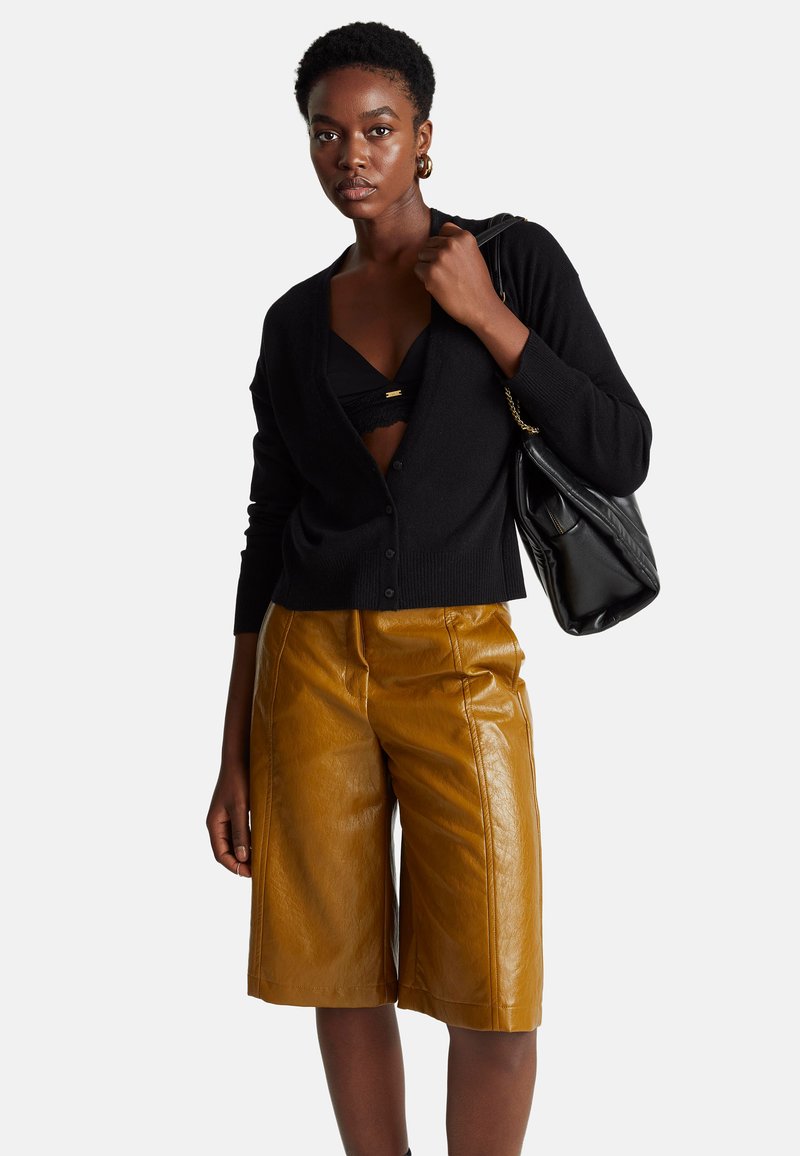 Czarny kardigan zapinany na guziki, połączony z czarnym koronkowym biustonoszem i beżowymi skórzanymi szortami wysokiego stanu. Trzyma dużą czarną torebkę.