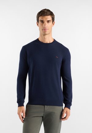 Uomo che indossa un maglione blu navy a maniche lunghe e pantaloni verde oliva, in piedi davanti a uno sfondo chiaro e uniforme.
