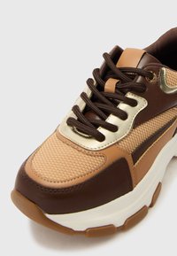 Brune og tan snøre med mesh-overdel, guldfarvede detaljer og brune snørebånd. Udstyret med en kraftig hvid sål og strukturerede detaljer på siderne.
