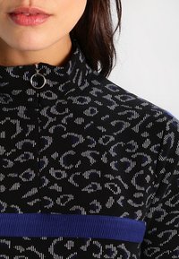 Top negro con un estampado, de cuello alto, que presenta una cremallera y una franja horizontal azul. La tela tiene un diseño de estampado animal texturizado.