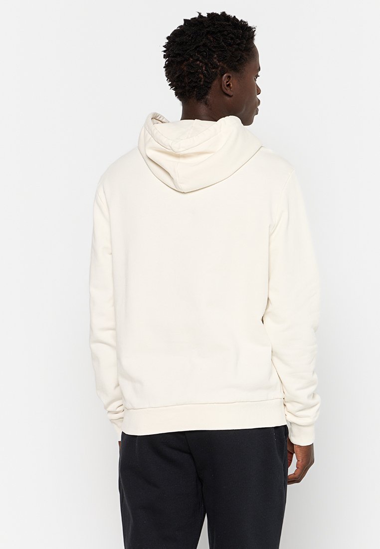 Uomo con capelli corti e ricci che indossa una felpa bianca con cappuccio e pantaloni neri, di spalle contro uno sfondo bianco uniforme.