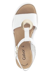 White leather sandal with a round gold hardware accent, adjustable strap, and cushioned footbed. Flat sole with a beige color.