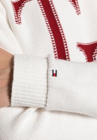 Pull blanc en tricot avec un grand logo rouge, des surpiqûres accentuées et un poignet côtelé orné de petits détails bleu marine, rouge et blanc.