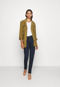 Chaqueta ligera verde oliva con mangas dobladas, dos bolsillos frontales y botones, combinada con una blusa blanca, jeans azul oscuro y tacones amarillos.