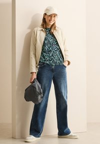 Veste bomber beige clair, haut à motifs turquoise, jean large bleu foncé, baskets blanches, tenant un sac gris, debout contre un mur clair.