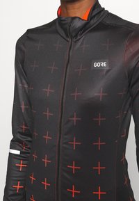 Sort cykeltrøje med rødt geometrisk krydsmønster, lynlås foran, høj krave og GORE-logo. Glat stof med en tætsiddende design.